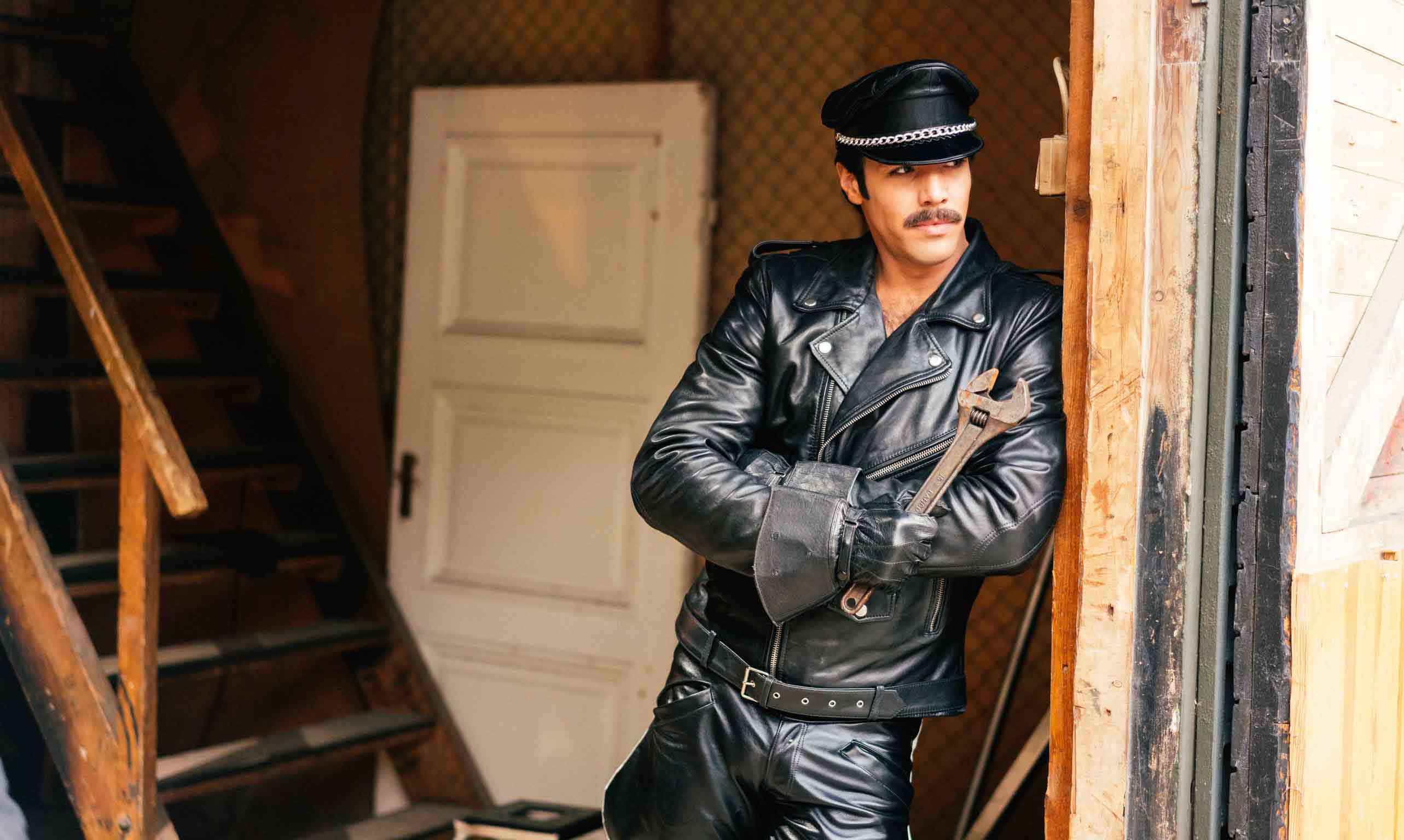 Från Tom of Finland, Finlands bidrag till Oscarsgalan. En man i läderkläder står lutad mot en vägg med en stor skiftnyckel i handen. 