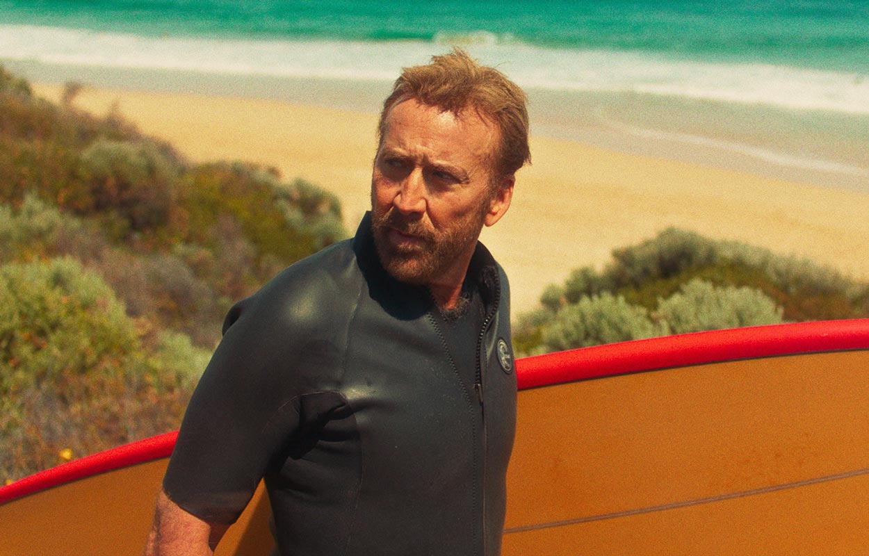 Nicolas Cage står vid stranden med en surfbräda. 