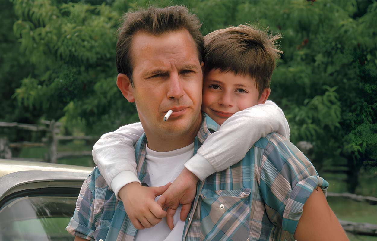 Kevin Costner röker en cigarett och har en pojke på sina axlar.